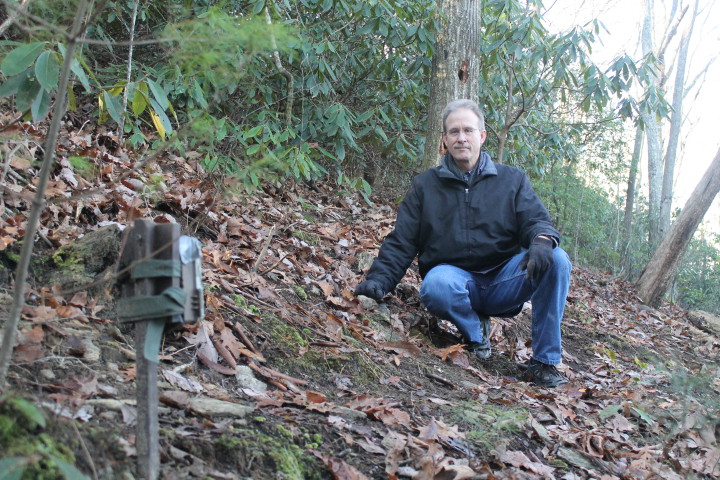 Tom Brass kneels at one of his three trail camera sites behind his Henderson County home, where he has captured videos of bears, bobcats, coyotes, foxes and more. (Photo by Rachel Ingram).