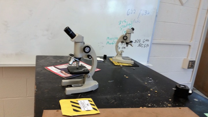 Microscopes sit on a desk in a science classroom.